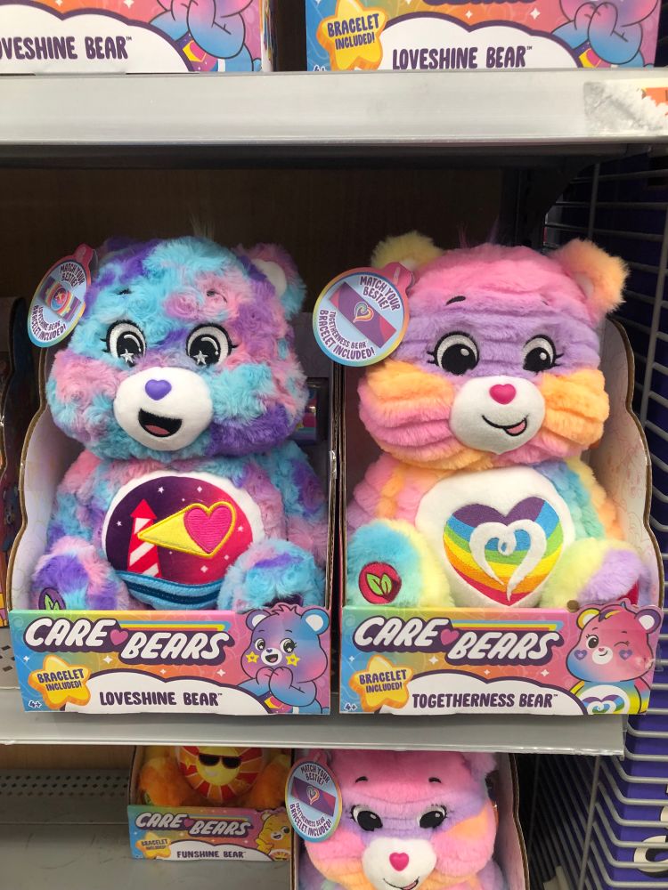 Two stuffed Care Bears toys on a store shelf. One is blue, purple, and pink with a lighthouse on his belly. He is called Loveshine Bear. The other one is multicolored with a rainbow heart on his belly. His name is Togetherness Bear.