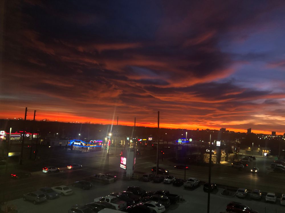 A heavy layer of dark clouds cover the top of the picture illuminated in orange and blue and purple as the sun sets low in the sky behind a dark city skyline. There is a swath of bright orange below the clouds. In more immediate view is a parking lot and several fast food restaurants.