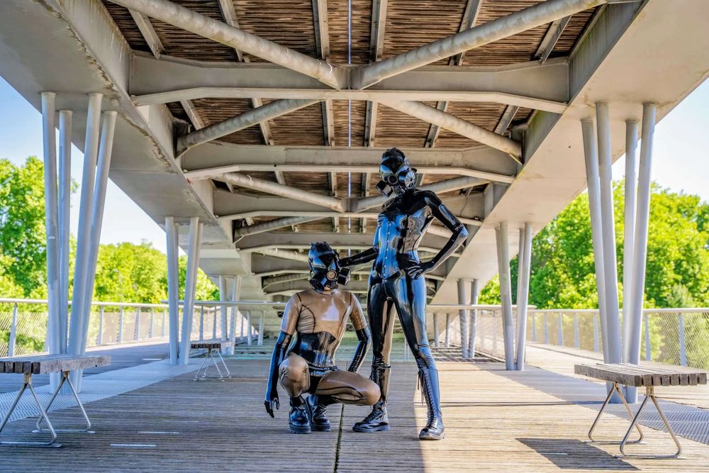 Two latex drones stands confidently on a modern steel bridge. The architectural lines of the bridge create a futuristic backdrop, echoing the sleek and bold aesthetic of the outfit. The scene is bathed in daylight, highlighting the contrast between industrial structure and avant-garde fashion.