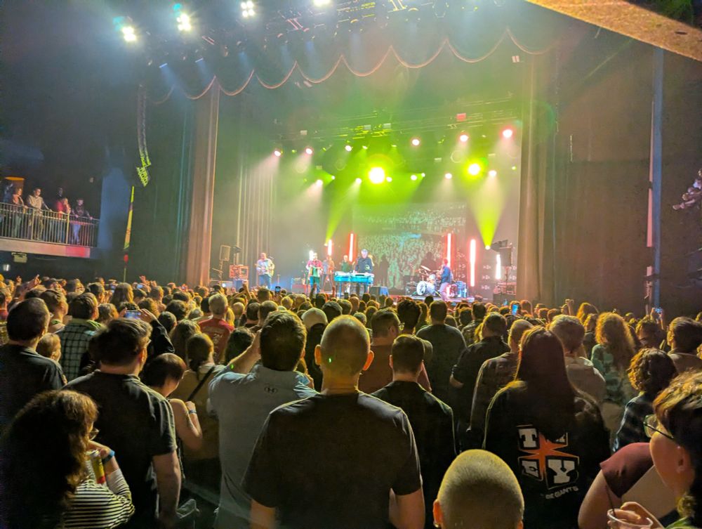 A crowd before the colorful lit and smokey stage at a TMBG show