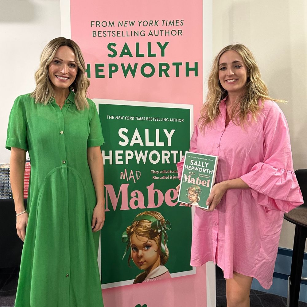 Sally and Amy smile for a photo in front of a pull-up banner for Mad Mabel, which features a pink background, green text that says 'From New York Times bestselling author Sally Hepworth', the Pan Macmillan Australia logo, and an image of the front cover of Mad Mabel, which has a green background, the book title and a graphic of a young girl.