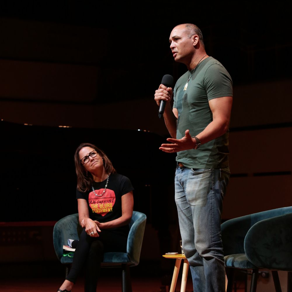 Thomas is shown standing and speaking while Clare Wright sits beside him, listening.