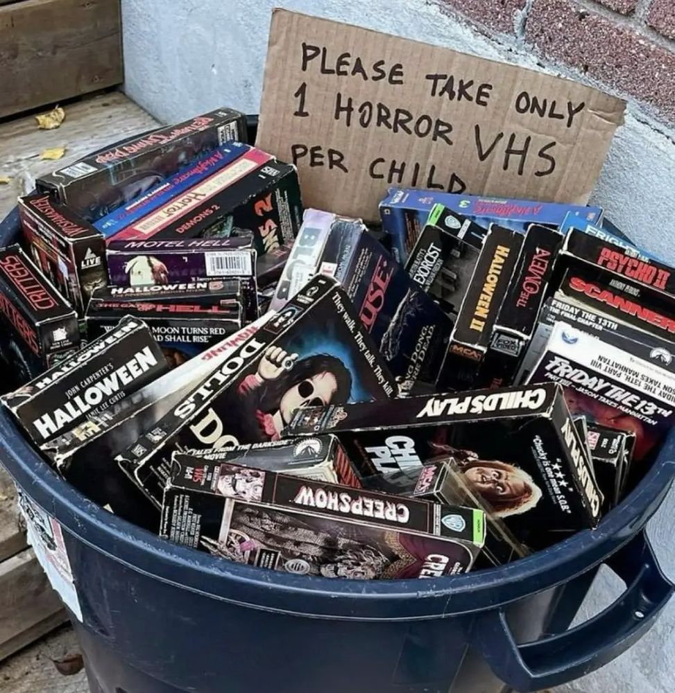 A bucket of VHS tapes of horror movies with a sign that says, "Please only take one horror VHS per child." 
