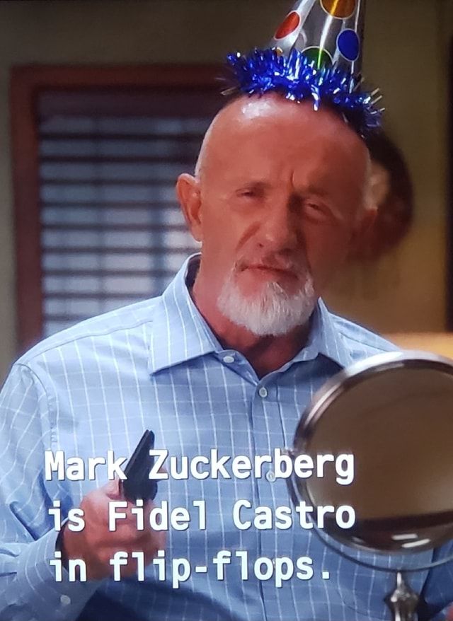 Buzz Hickey, a bald man with a white goatee wearing a blue button-up shirt and a party hat, stands in front of a small mirror in his office. Text in front of him reads "Mark Zuckerberg is Fidel Castro in flip-flops."