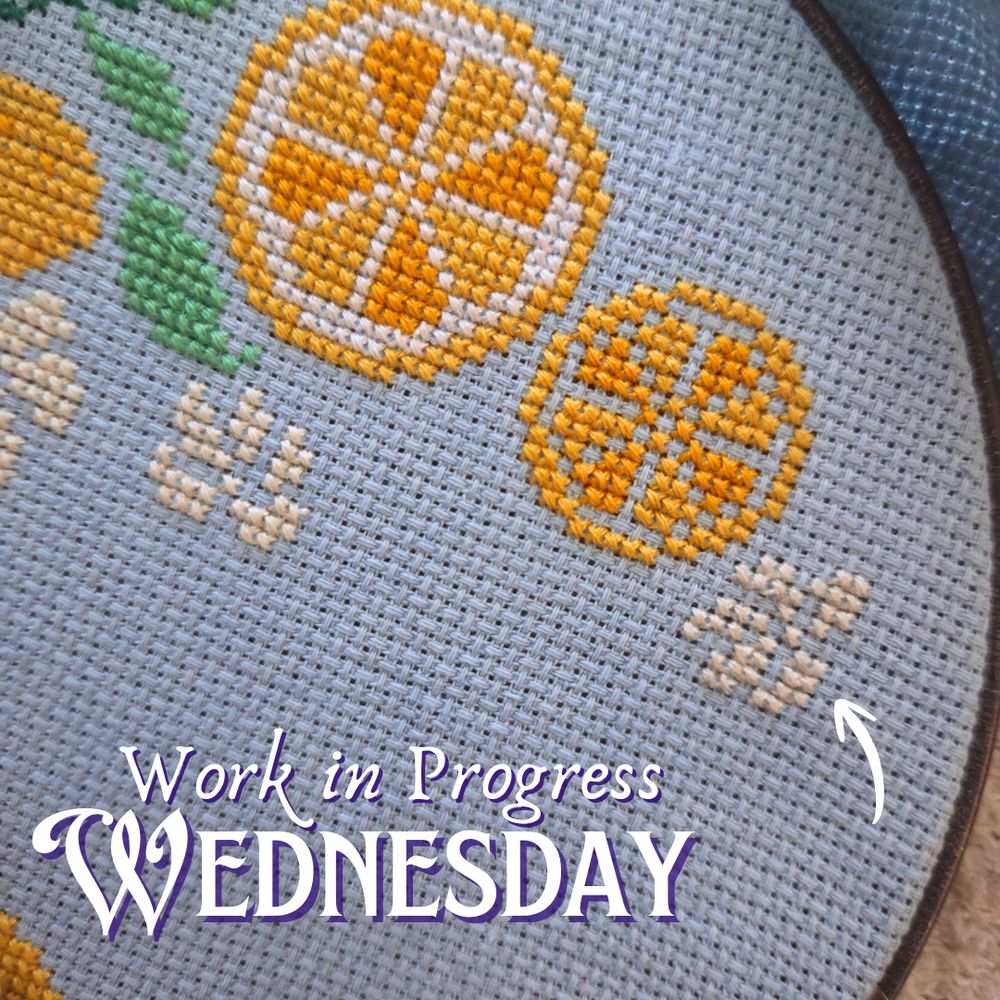 A close up of a cross stitch project in the works on light blue fabric, showing stitched slices of lemons, some green leaves, and some white flowers. Work in Progress Wednesday. A small arrow is pointing at a flower motif on the right hand side. 