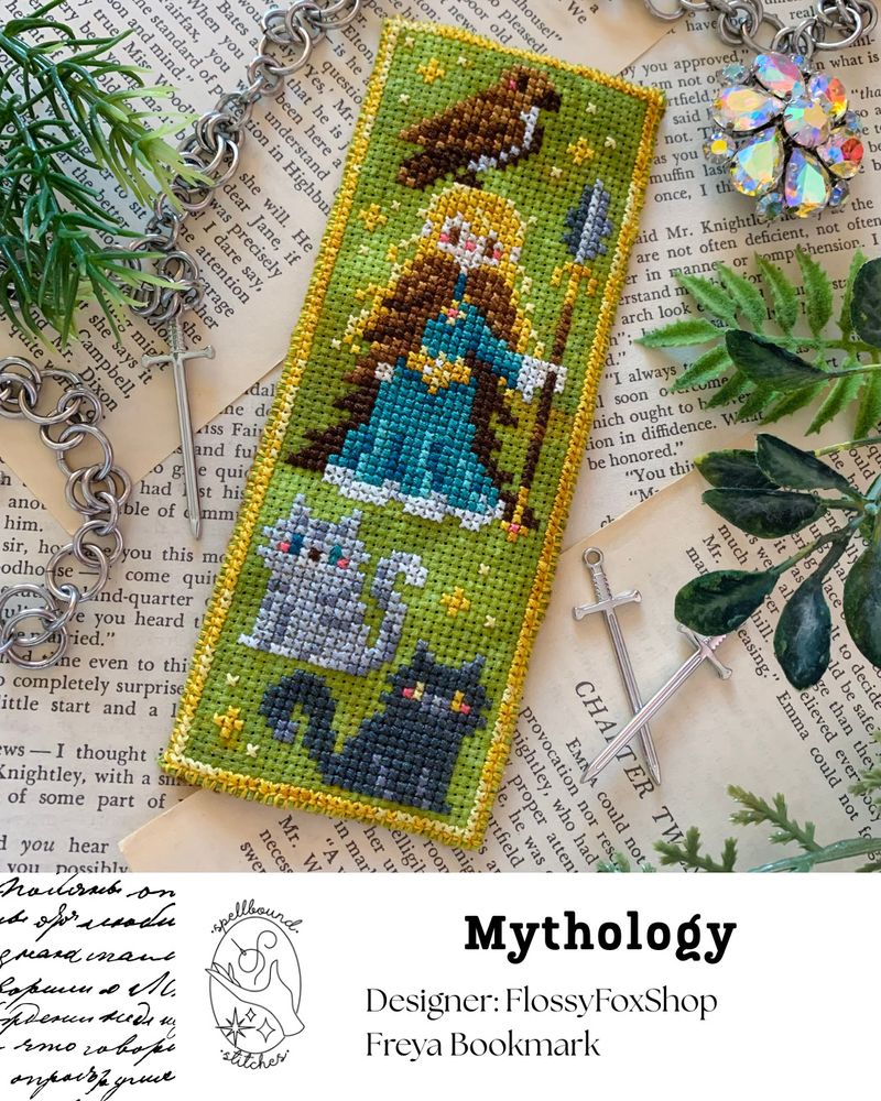 A cross stitch bookmark on moss green fabric showing Freya of Norse Mythology, with a feather cloak and a hawk perched upon her head. She is brandishing a spear and her two cats are sitting in front of her. Spellbound Stitches logo. Mythology. Designer FlossyFoxShop. Freya Bookmark.