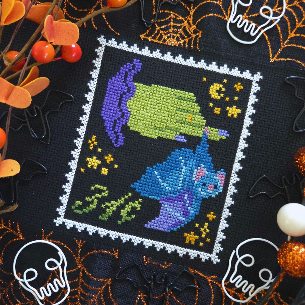 A staged cross stitch project stitched on black fabric showing a postage stamp with a witchy hand and a blue bat hanging upside-down from a finger. There's some starry motifs and 31 cents. 