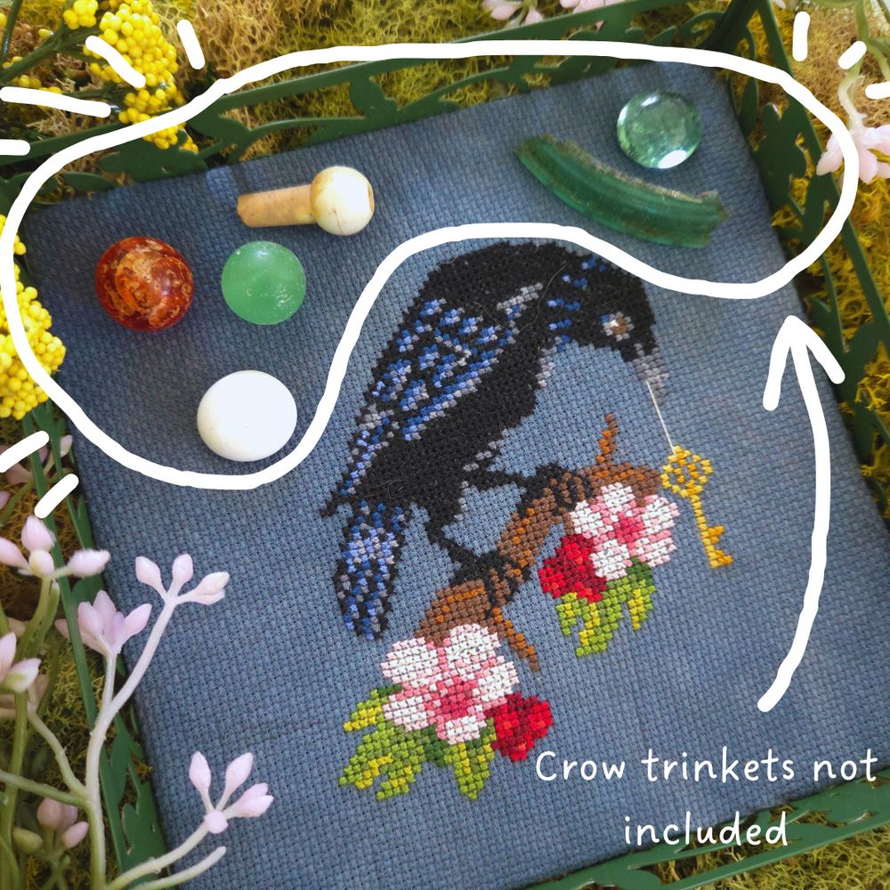 A cross stitch project finished in a green, floral tray and resting on a bed of moss.  The cross stitch shows a crow, perched on a blooming hawthorn branch, with a strung key hanging from their beak.  There are small trinkets on the tray, marbles, bits of shiny plastic with the notation, Crow Trinkets not included.