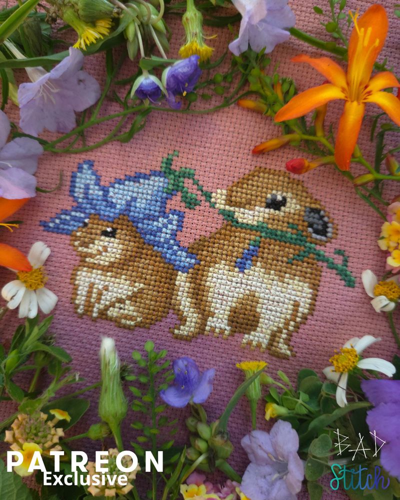 A cross stitch project staged with real wildflowers of varying colors and textures.  The projects shows a large pika with a stem in her mouth. The end of the stem leads to a downward facing bell flower, that is perched upon the head of a piklet (baby pika).  Patreon Exclusive. BAD Stitch.