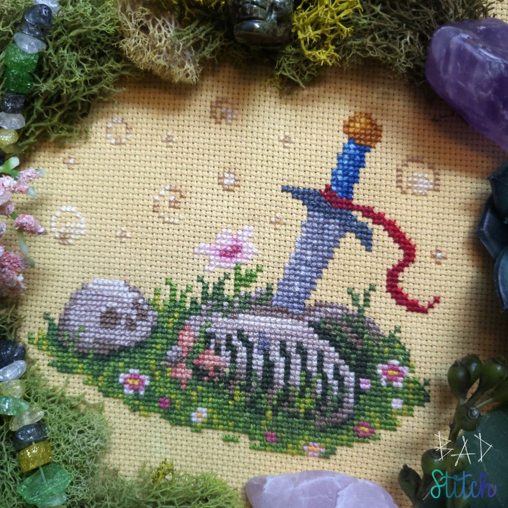 A cross stitch of a skull and ribcage, overcome with grasses, flowers, and mushrooms.  An abandoned sword is thrust into the chest of the ribcage with a red ribbon tied on the hilt, waving in the breeze. BAD Stitch.