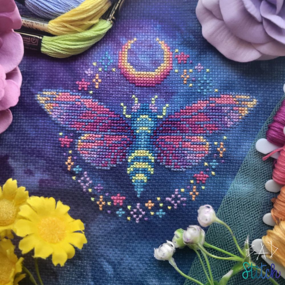 A cross stitch project of an open-wing cicada, surrounded by small star and moon motifs.  BAD Stitch