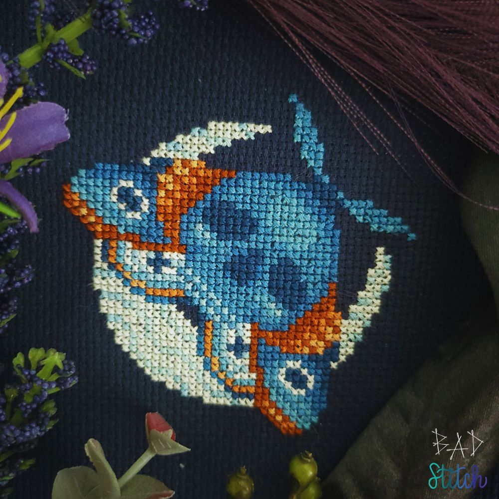A completed cross stitch project stitched on black fabric depicting a large, stylized death-head moth flying over a crescent moon.  BAD Stitch.