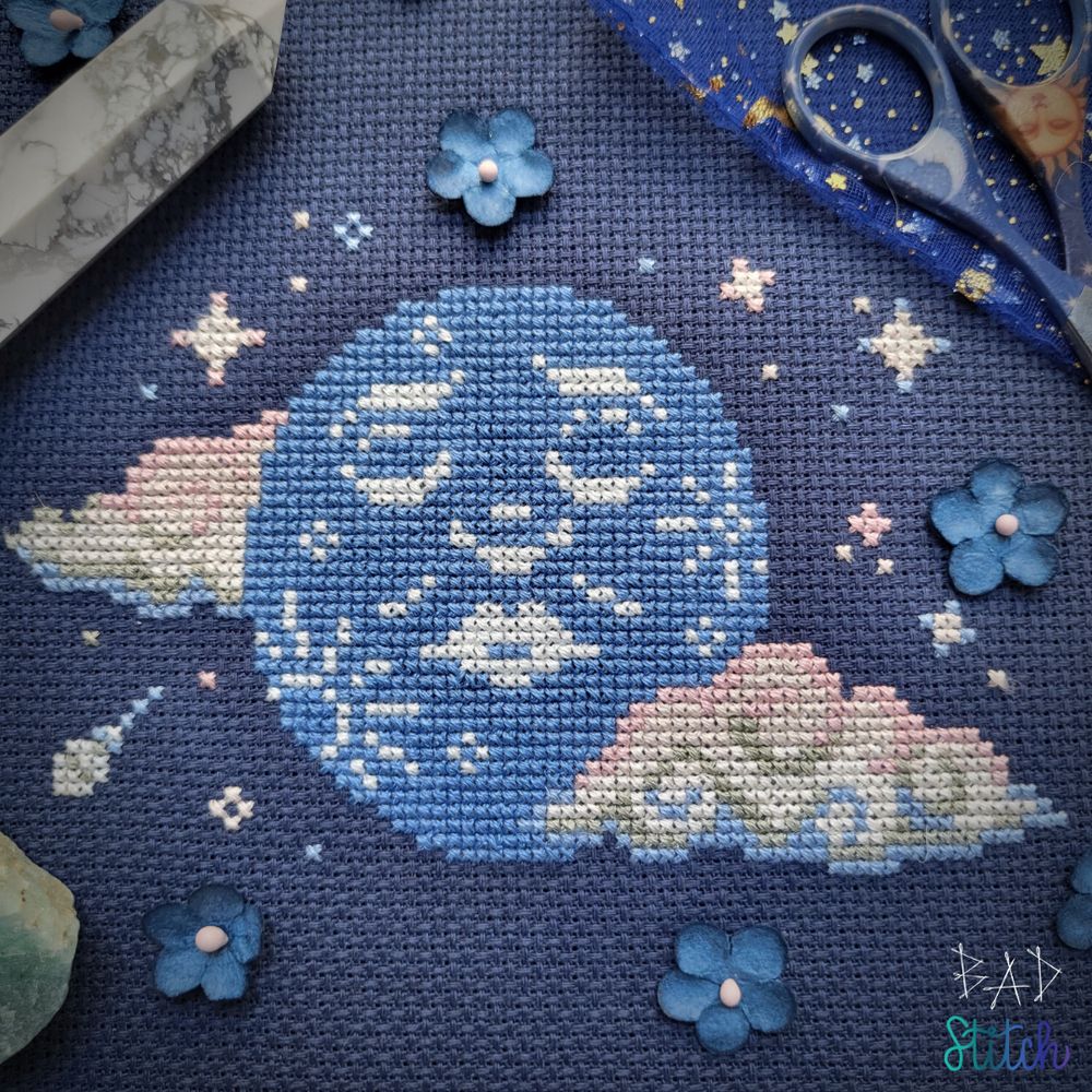 A staged cross stitch project on navy fabric showing a big blue moon with a somber face. Big clouds and starry bits surround the moon. BAD Stitch.