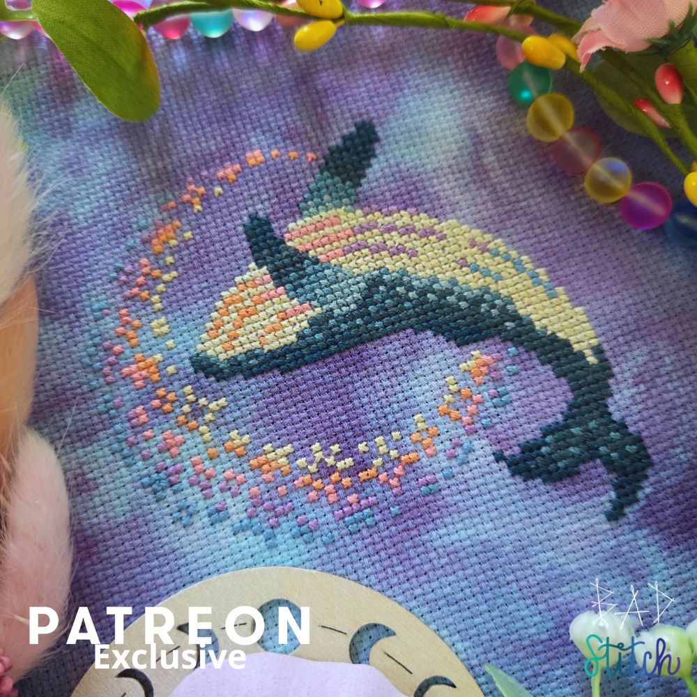 A cross stitch project on mottled fabric and staged with fake florals, translucent beads, and a wooden moon shape. The cross stitch depicts a humpback whale, jumping backwards into a moon shape made of small starry motifs.