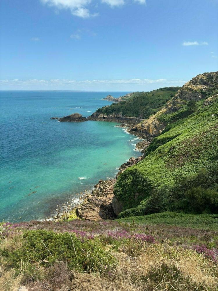 A coastal landscape features clear turquoise water, rocky cliffs, and lush greenery under a partly cloudy sky.