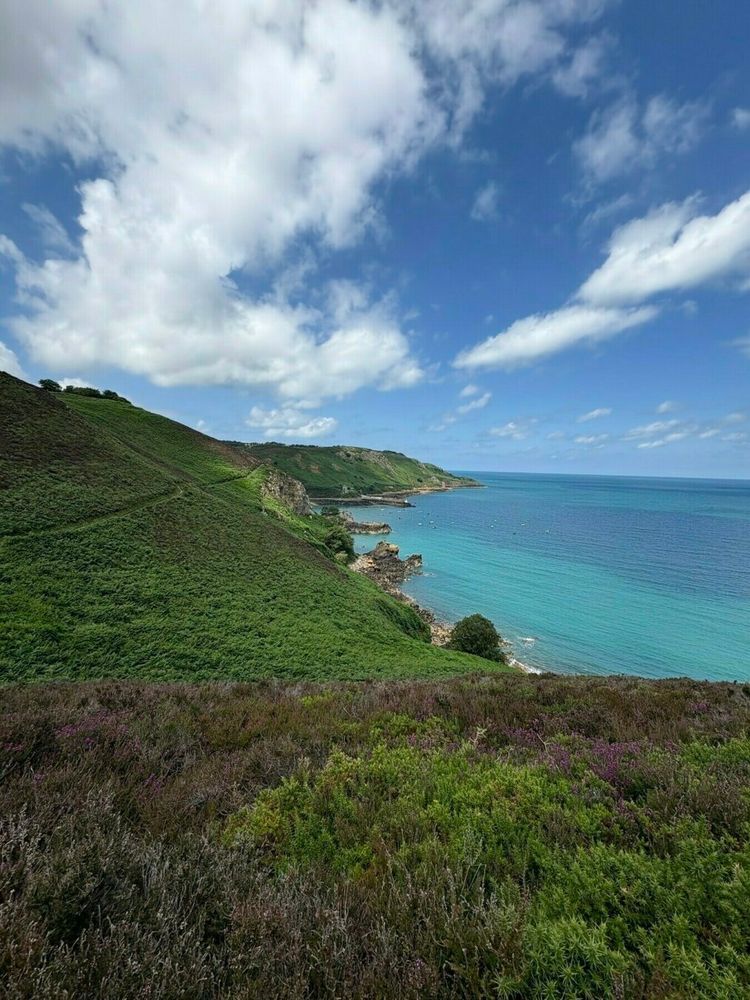 A scenic coastal landscape features a clear blue sea, rolling green hills, and a partly cloudy sky.