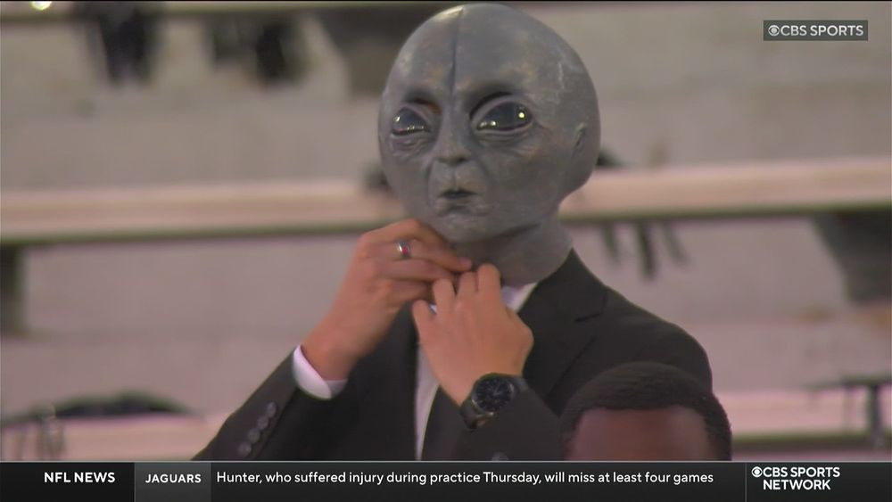 Dude at a college football game presumably dressed like a “men in black,” wearing a black suit & tie with an alien mask on his head. 