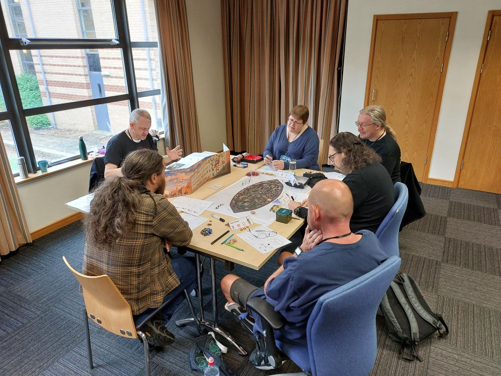 Table of roleplayers playing RuneQuest. Maps, standees and dice on the table. 
