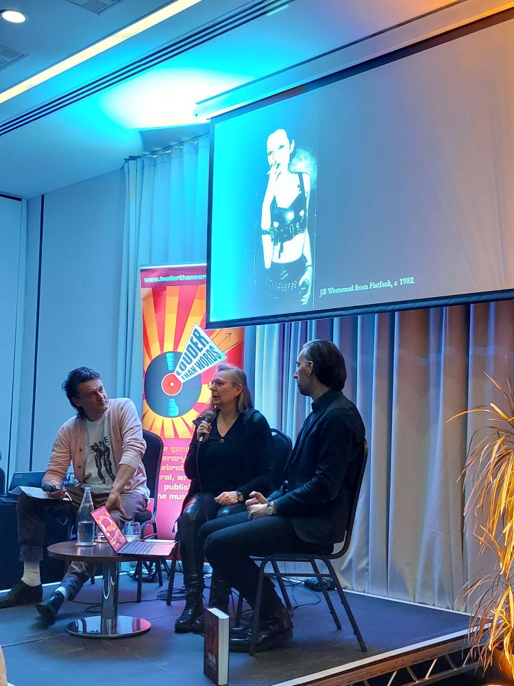Louder Than Words literary festival. Three speakers are seated on a small stage (L-R: Ian Trowell, Jill Westwood, Nicolas Ballet). The screen behind shows Jill in fetish gear, performing as part of Fistfuck, circa 1982.