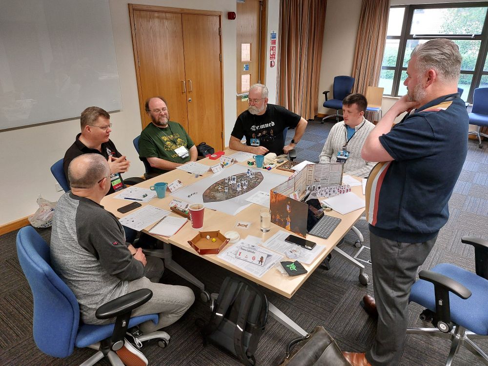 Table of roleplayers playing RuneQuest. Maps, standees and dice on the table. 