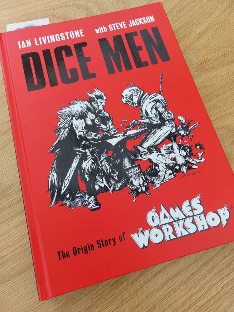 Book on a wooden table. Dice Men by Ian Livingstone with Steve Jackson. The origin story of Games Workshop. Plain red cover with bold title text, and a black and white picture of a fantasy warrior and a spaceman playing a table-top game.