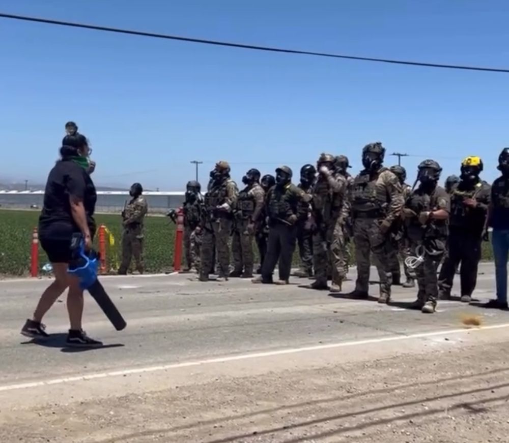 Person with air blower stands down in front of ICE troops with tactical gear. 