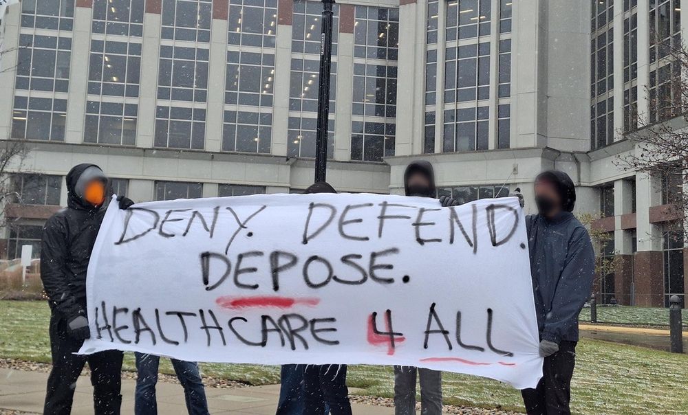 Protesters in front of medical building hold banner reading, "Deny, Defend, Depose, Healthcare 4 All."