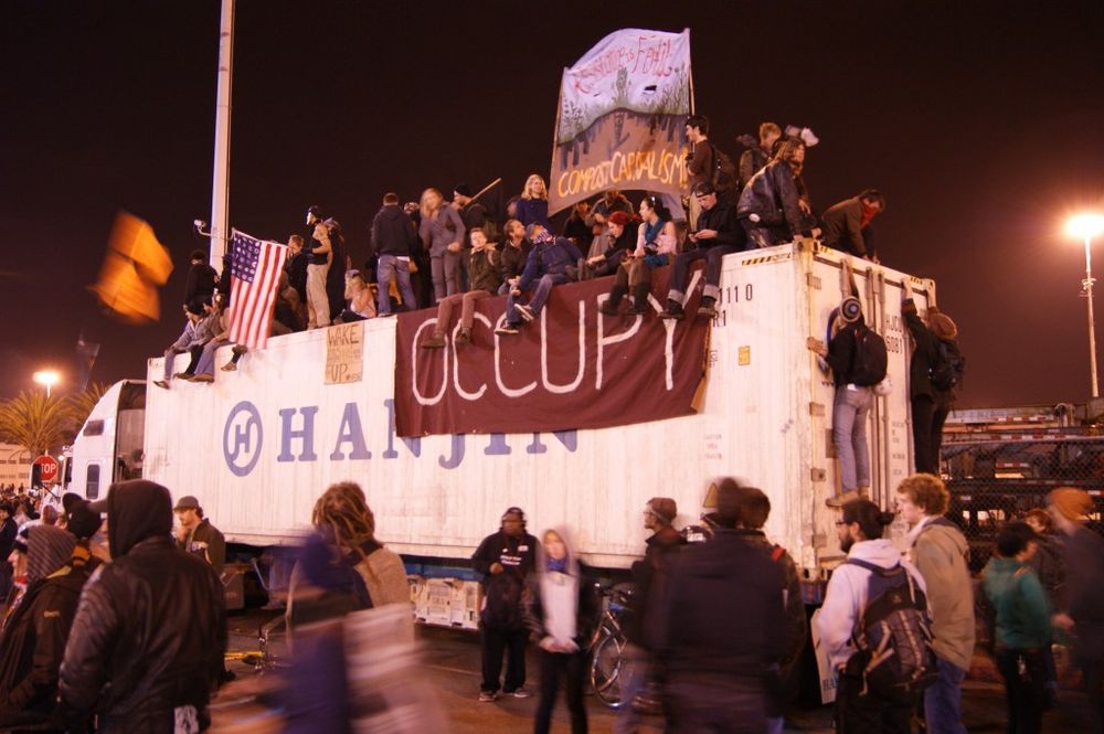 Photo from Occupy Oakland port shutdown in 2011. People on shipping container with the word, "Occupy" on a banner. 