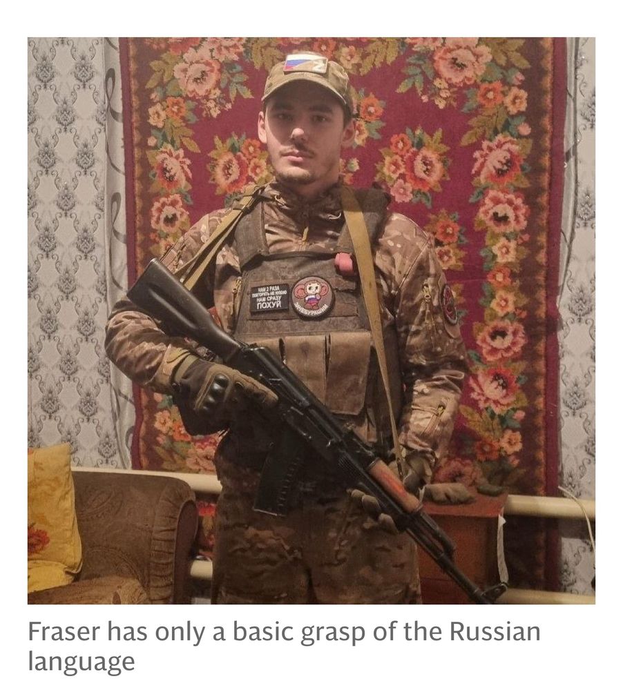 Picture of Jay Fraser, from Scotland, standing in front of a hanging rug and wearing the uniform of some tin pot Russian PMC. He has a Kalashnikov pattern rifle. He is a moron. Fuck him. Caption: Fraser has only a basic grasp of the Russian language. 