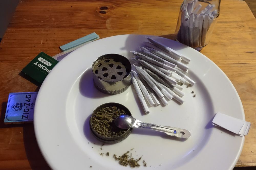 a table with a plate and jar on it, along with a grinder, a small spoon, some packets of rolling papers, some small pieces of card for folding filters, and 56 freshly rolled joints (some on the plate, some in the jar)