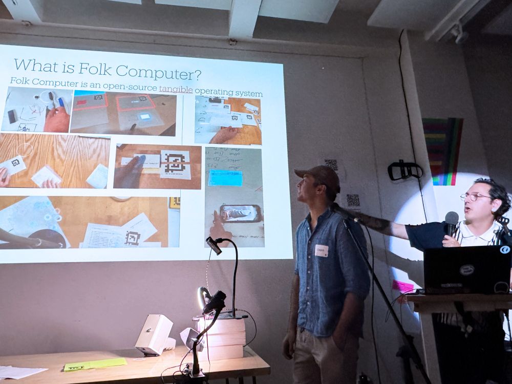 Andrés and Omar at a podium presenting a slide with many pictures that says "What is Folk Computer?"