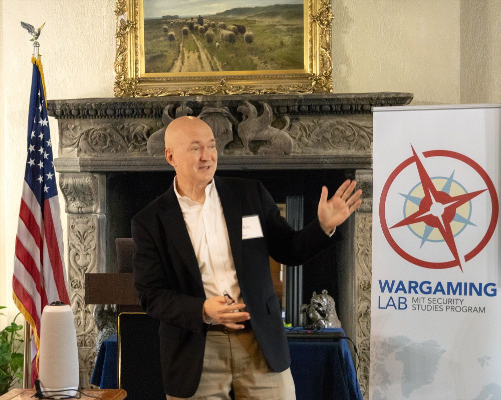 Eric Heginbotham lectures in front of a fireplace and American flag.
