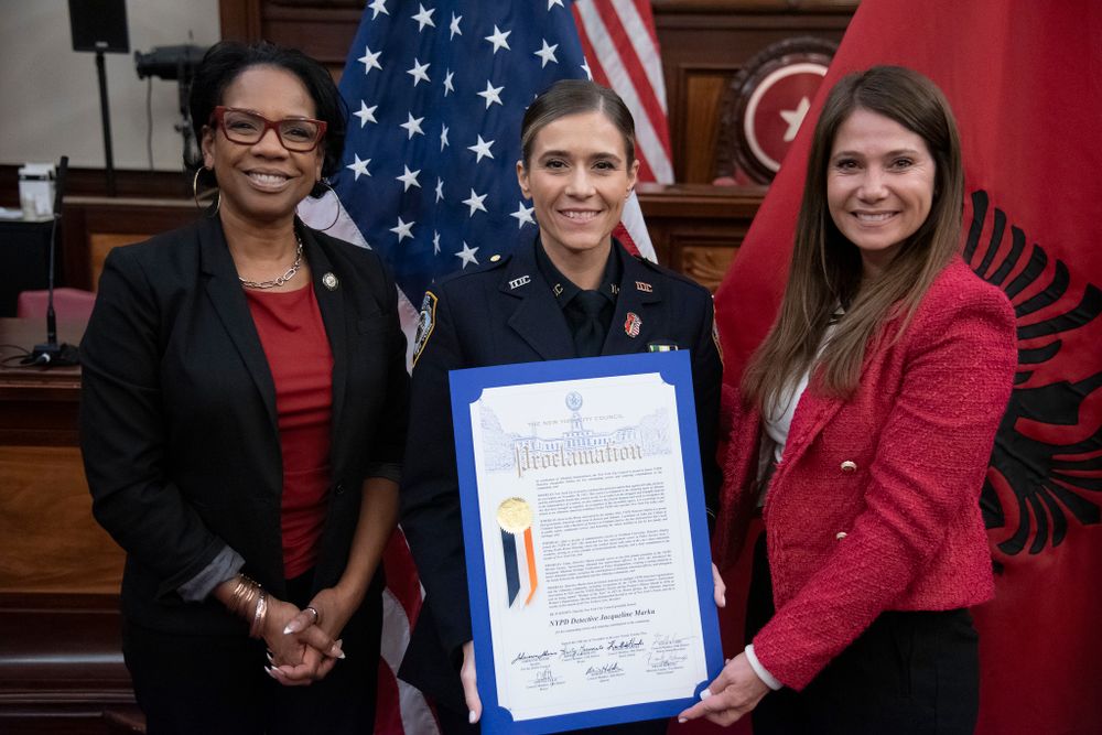 Council Members Kamillah Hanks and Kristy Marmorato hold a proclamation with a police officer for an Albanian Heritage celebration in Council Chambers. 
