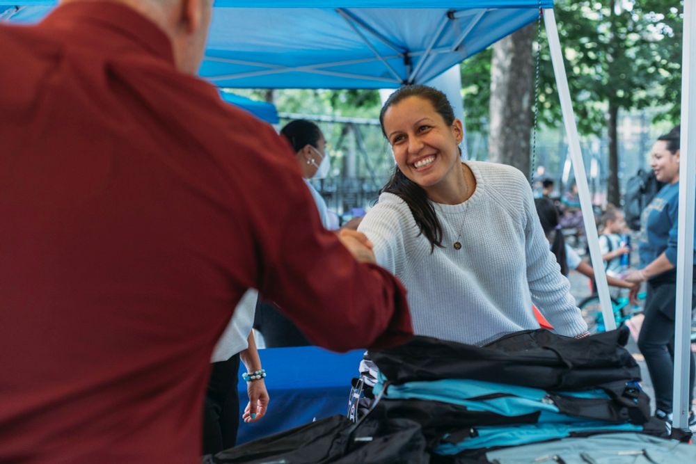 Councilmember Tiffany Caban smiling and shaking hands with another individual under a blue canopy at an outdoor event.