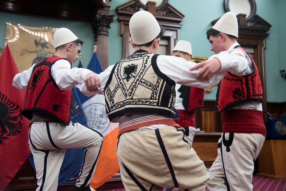 Three men dressed in traditional Albanian folk costumes perform a circle dance indoors with national flags in the background.