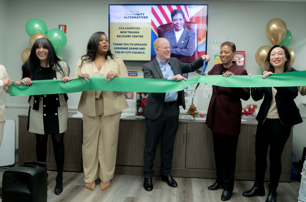 Several elected officials and community leaders hold a long green ribbon as it is cut during the ribbon cutting ceremony, with Speaker Adrienne Adams in a red blazer near the center.