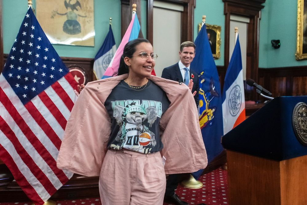 Council Member Tiffany Cabán showing off her Ellie the Elephant NY Liberty t-shirt with Council Member Erik Bottcher smiling in the background.