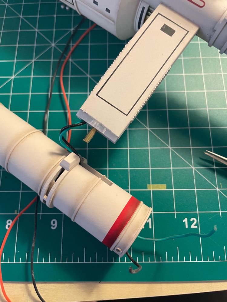 A Daedalus class model’s engine and strut sit on a green hobby mat. A brass support rod and a set of green and black wires jut from the support. The wires are fed through the engine and out it’s front section.