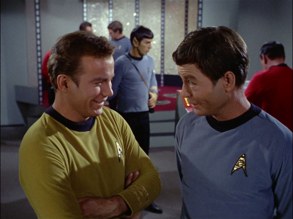 McCoy jokes with Kirk as Spock and other crew men work in the background of the transporter room. From the episode “Arena”.