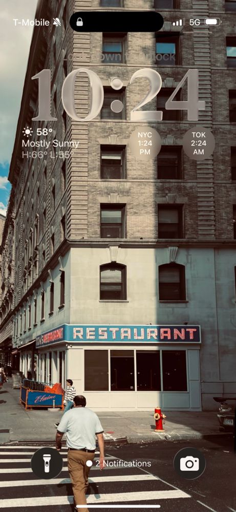 A photo of the outside of Tom’s Restaurant in Upper Manhattan on a sunny afternoon. The view is from across w 112th street on the east side of broadway. A man in brown pants is on the crosswalk. 