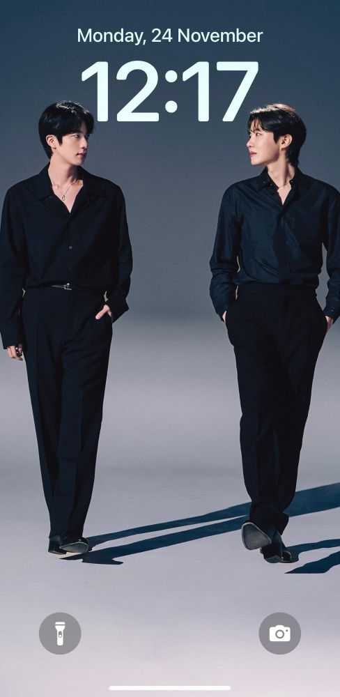 Jin and j-hope of BTS in black shirts and pants, walking parallel as they turn their heads to look at each other