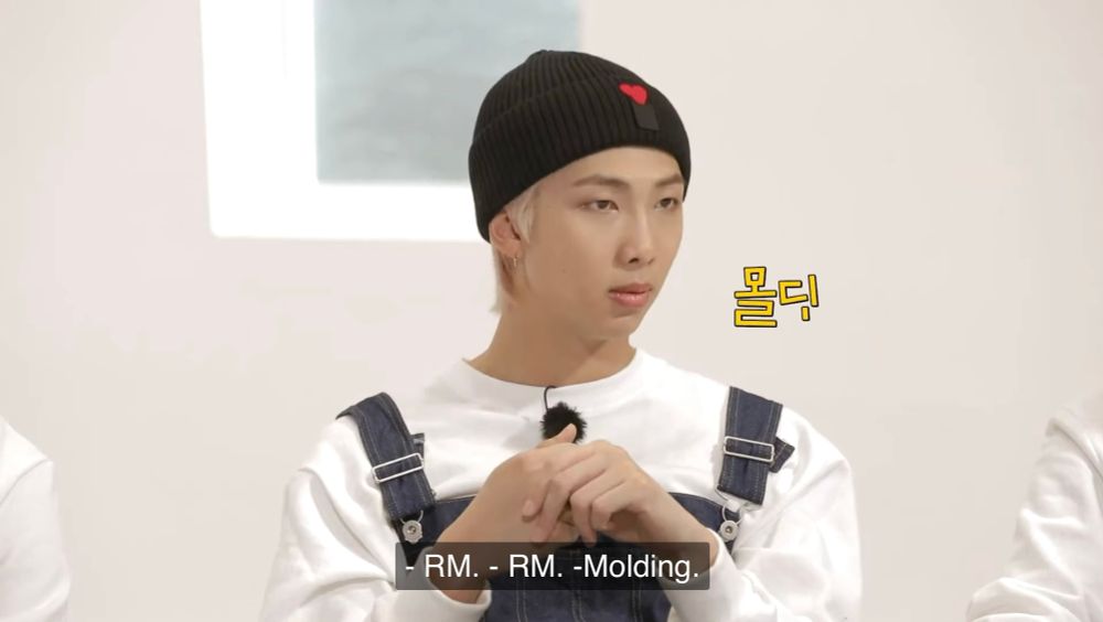 RM of BTS, in a white, long sleeved sweater and denim overalls, answers “Molding” to a quiz question
