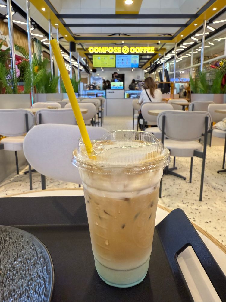 A photo of coffee with cream foam, in a plastic cup with straw. It sits on a black tray, in a cafe.