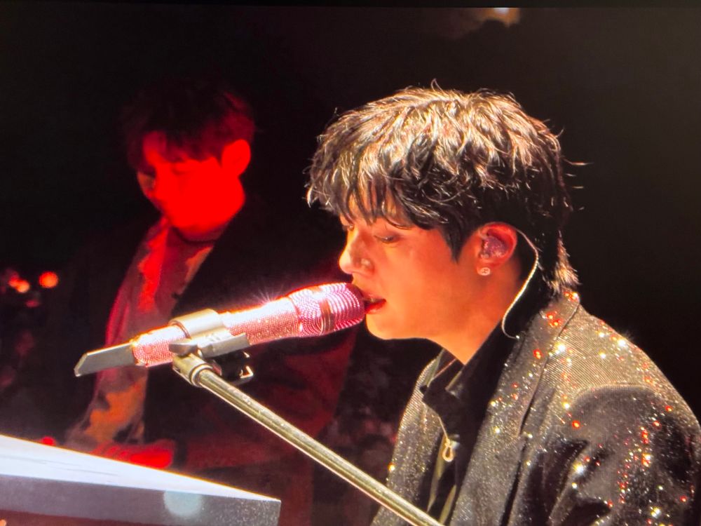 BTS’ Taehyung and Jin, who is singing into a mic as he plays a piano 