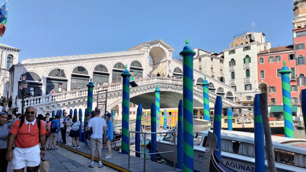 Blick vom Ufer des Canal Grande auf die Rialto-Brücke. Im Vordergrund sieht man außerdem blau-grün geringelte Pfosten, an denen Gondeln festmachen können. 