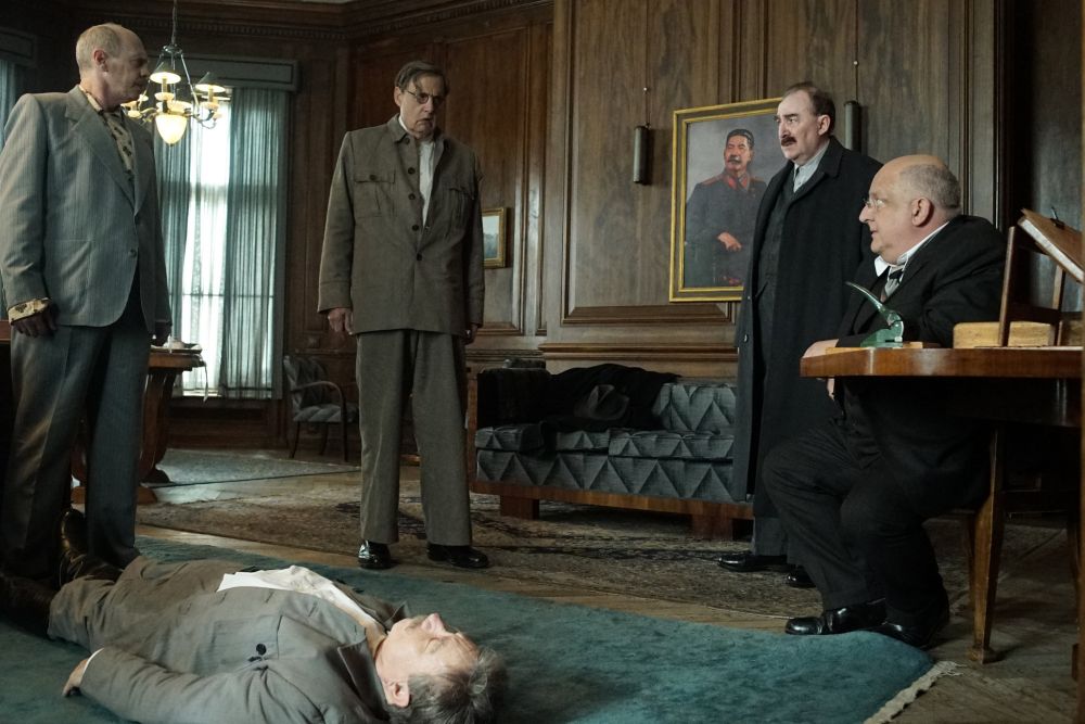 Screencap from The Death of Stalin. Khruschev (Steve Buscemi), Malenkov (Jeffrey Tambor), Kaganovich (Dermot Crowley), and Beria (Simon Russell Beale), standing dazed around Stalin's dead body in his private office. 