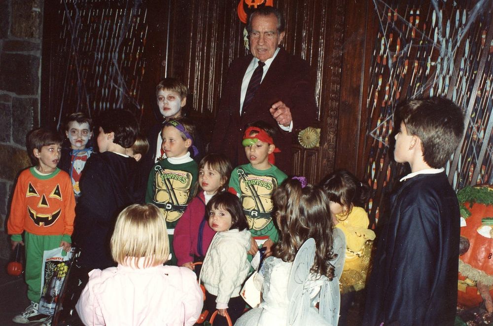 An elderly Richard Nixon on Halloween surrounded by trick or treating children in costume, including two in Teenage Mutant Ninja Turtles outfits. 