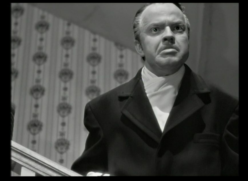 Citizen Kane screencap. Orson Welles as a middle-aged Charles Foster Kane, leaning over a banister, wearing a black overcoat and light scarf folded around his neck.