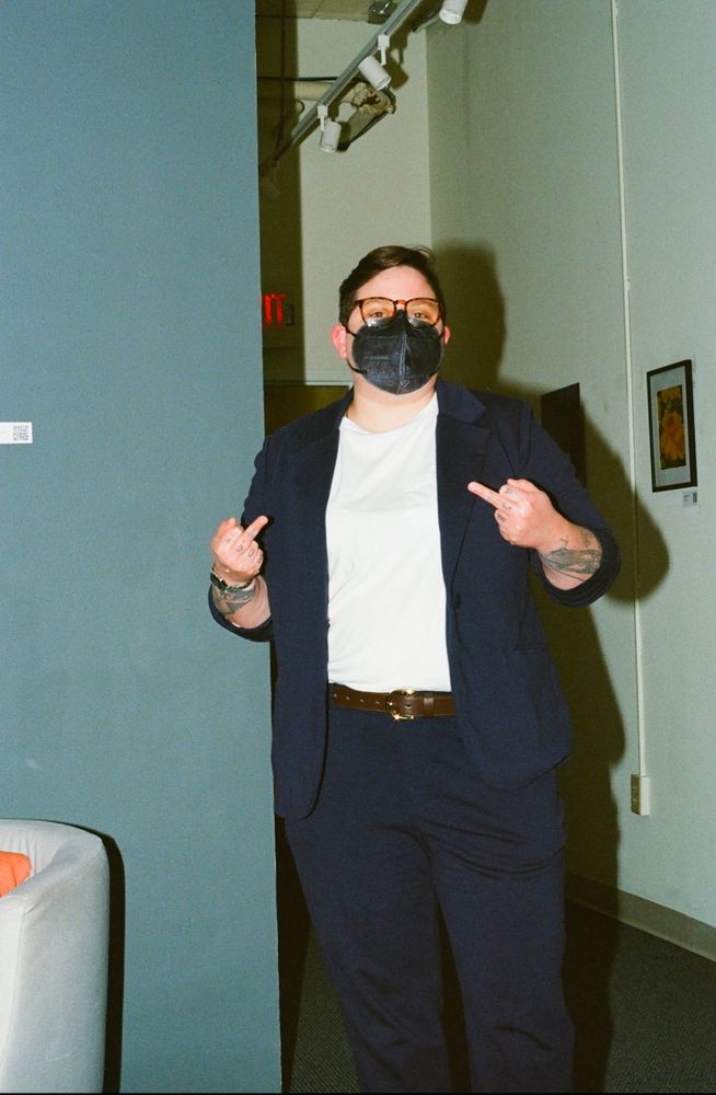 A white butch woman wearing a navy suit, white shirt, and a black mask flips off the camera. She is standing up