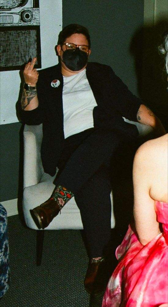 A white butch woman in a navy suit, white shirt, sits down on a white chair when flipping off the camera. There is a button on her blazer that says "Free Palestine".